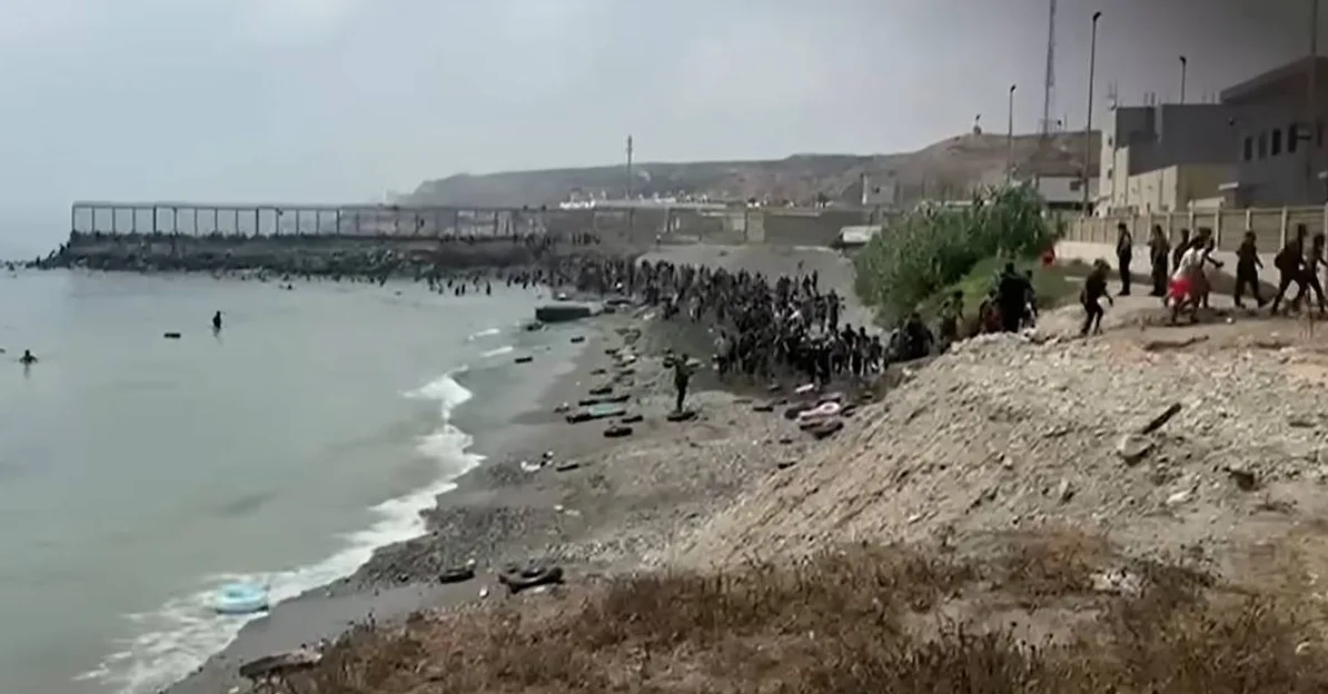 Velika grupa ljudi, uključujući djecu, okupljena je na šljunčanoj obali uz more. Neki ljudi plivaju u vodi, dok drugi stoje na obali ili hodaju po njoj. U pozadini se naziru građevine i ograda, sugerirajući graničnu ili infrastrukturnu zonu. Na obali su vidljivi odbačeni predmeti poput guma.