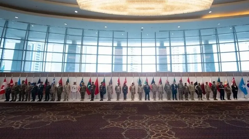 Grupa ljudi u vojnim uniformama pozira ispred niza zastava različitih zemalja.