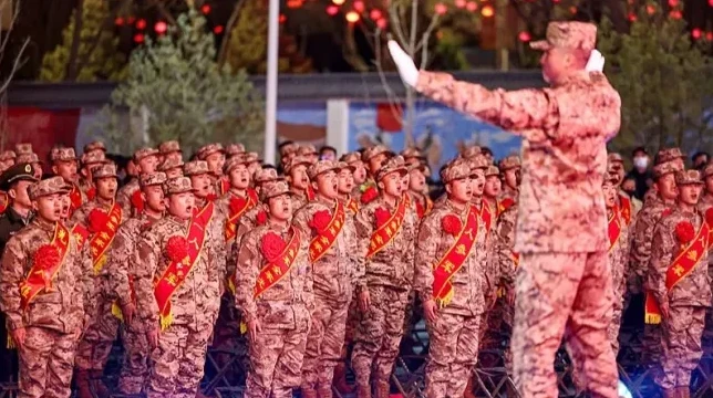 Grupa vojnika u maskirnim uniformama sa crvenim trakama preko grudi i ukrasima u obliku cvijeta, izvodi počasni manevar pod nadzorom oficira.