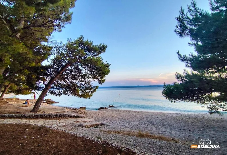 Plaža sa šljunkom i drvećem u prvom planu, sa pogledom na mirno more i blagi zalazak sunca u pozadini.