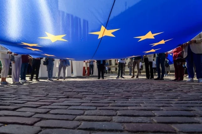 Grupa ljudi drži veliku zastavu Evropske unije iznad kaldrmisanog trga.