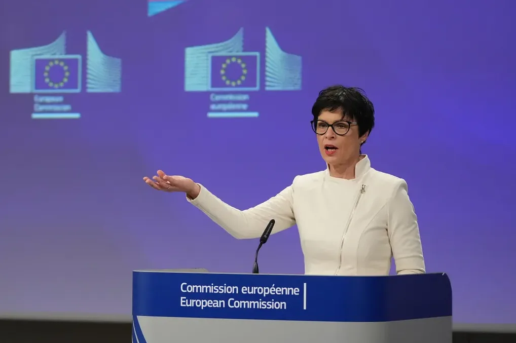 Žena sa kratkom crnom kosom i naočarima, obučena u bijeli sako, govori ispred postolja sa logom Evropske komisije.