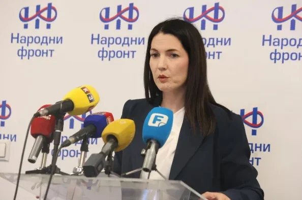 Žena s tamnom kosom govori iza postolja sa nekoliko mikrofona ispred transparenta sa logom 'Narodni front'