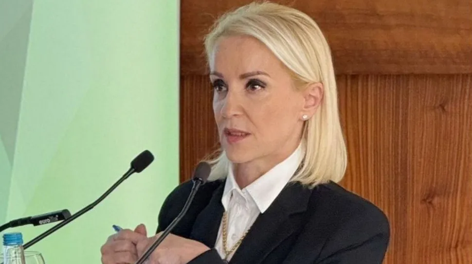 Plavokosa žena u crnom sakou i bijeloj košulji govori za govornicom ispred mikrofona.