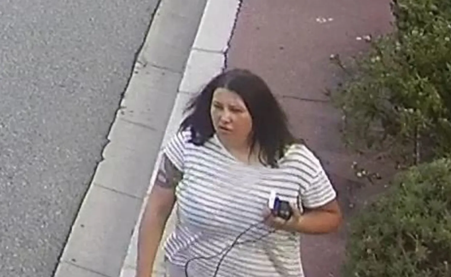 Žena sa tamnom kosom, nosi prugastu majicu, drži mobilni telefon u ruci, hodajući pored pješačke staze.