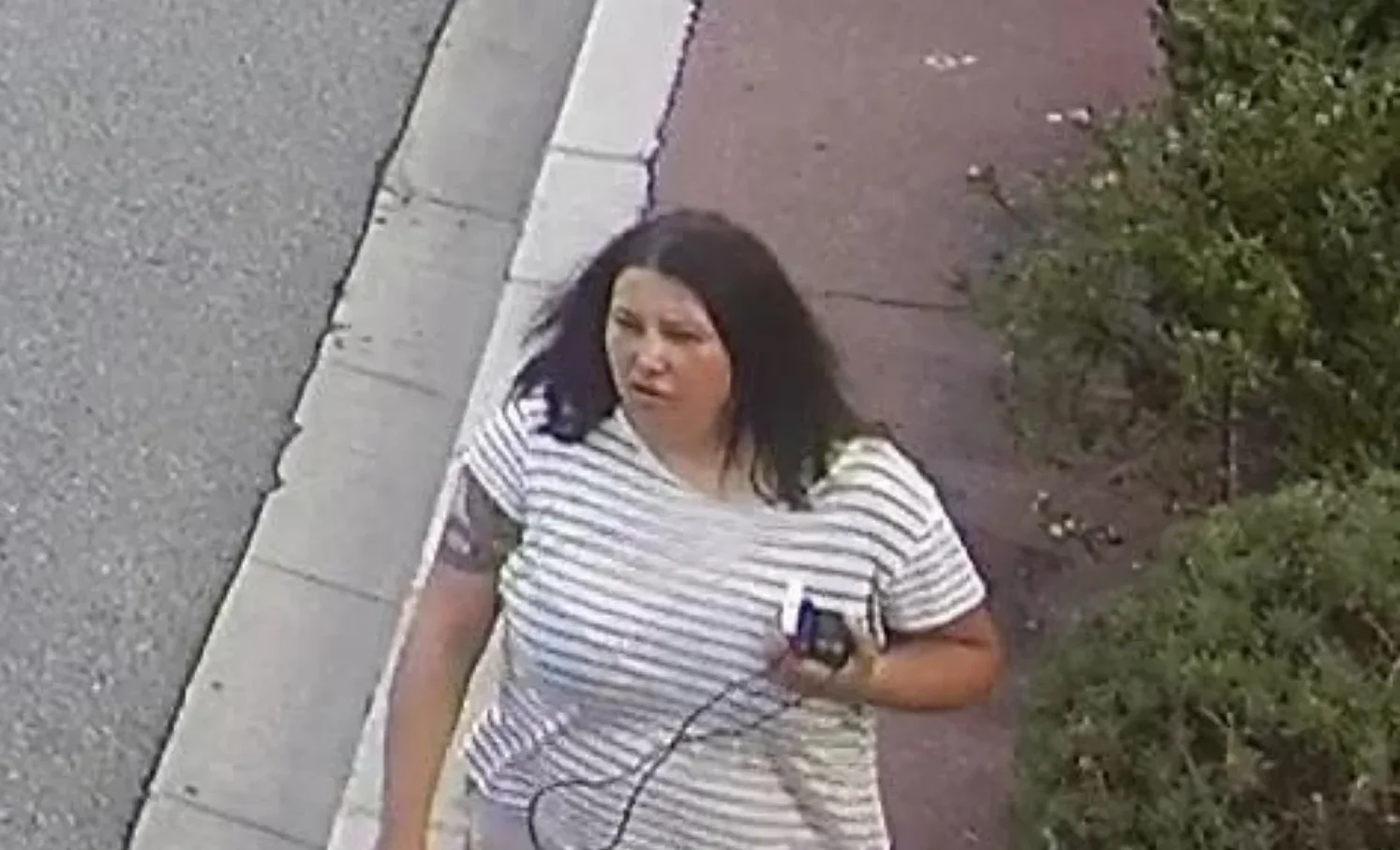 Žena sa tamnom kosom, odjevena u majicu na pruge, drži mobilni telefon u ruci dok stoji pored ograde i zelenila.