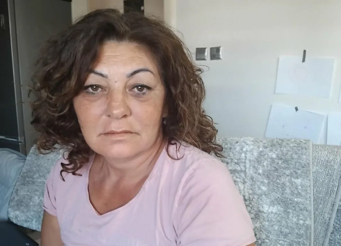 Žena srednjih godina sa smeđom kovrdžavom kosom i u roze majici sjedi na fotelji, gledajući ka kameri sa neutralnim izrazom lica.