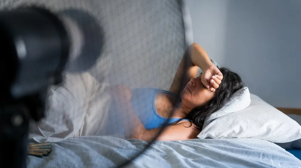 Žena leži u krevetu, iscrpljena, drži ruku na čelu i pati od vrućine dok je ventilator usmjeren prema njoj.