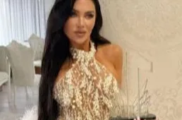 Žena u elegantnoj, prozirnoj haljini ukrašenoj biserima i perjem, drži nagradu.