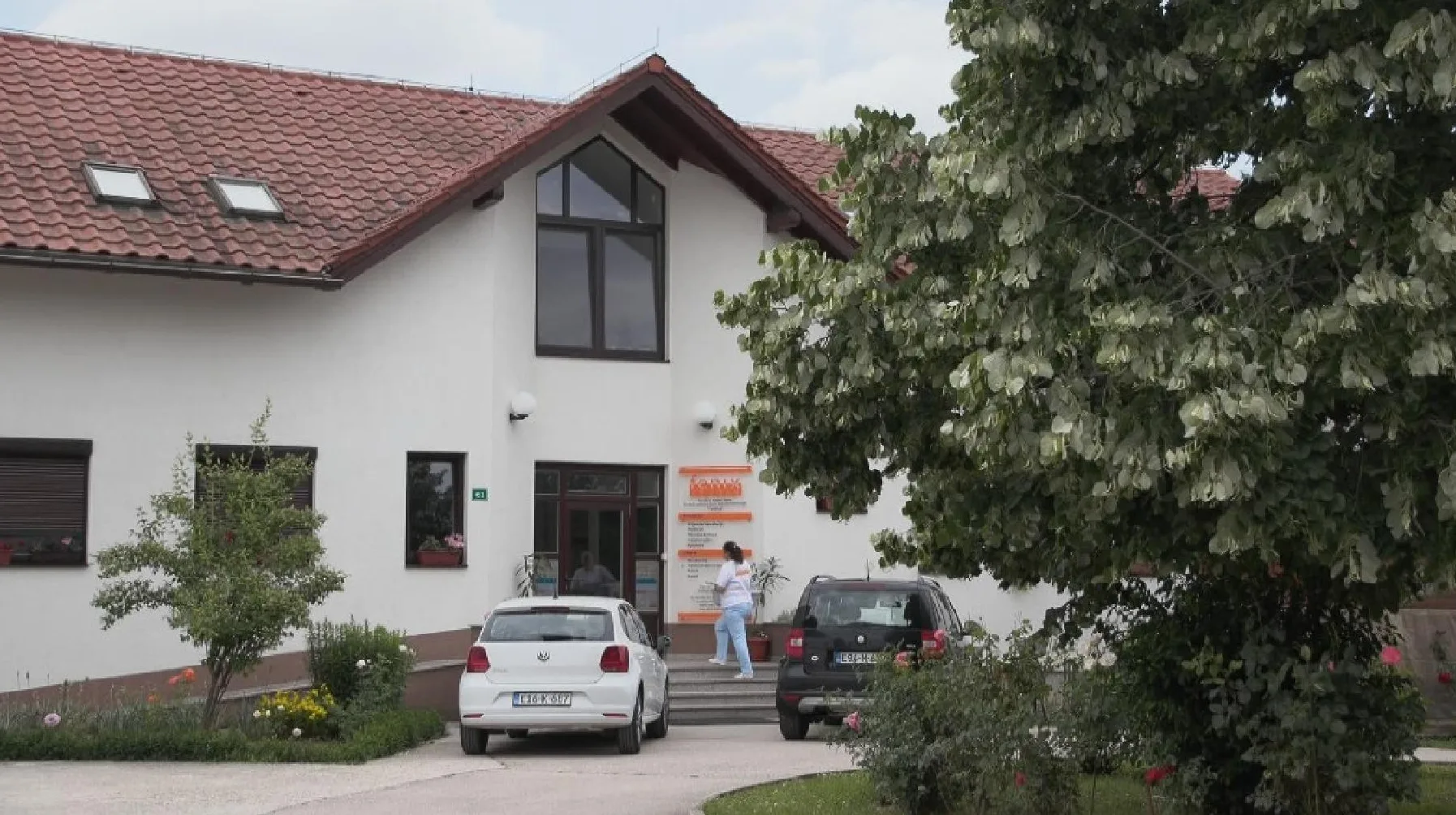 Bijela zgrada sa crvenim krovom, parkirani bijeli i crni automobil ispred, te veliko drvo sa zelenim lišćem sa desne strane.