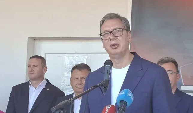 Aleksandar Vučić, predsjednik Srbije, govori sa mikrofonom, okružen drugim zvaničnicima.