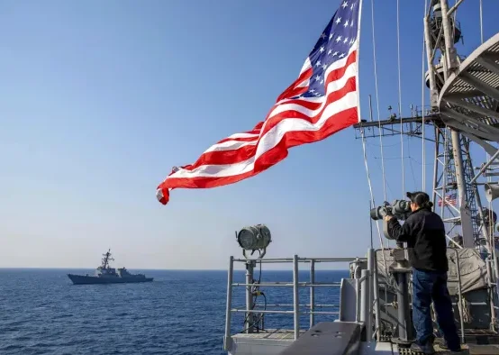 Američka zastava vijori na jarbolu vojnog broda, dok mornar promatra pomoću dalekozora.