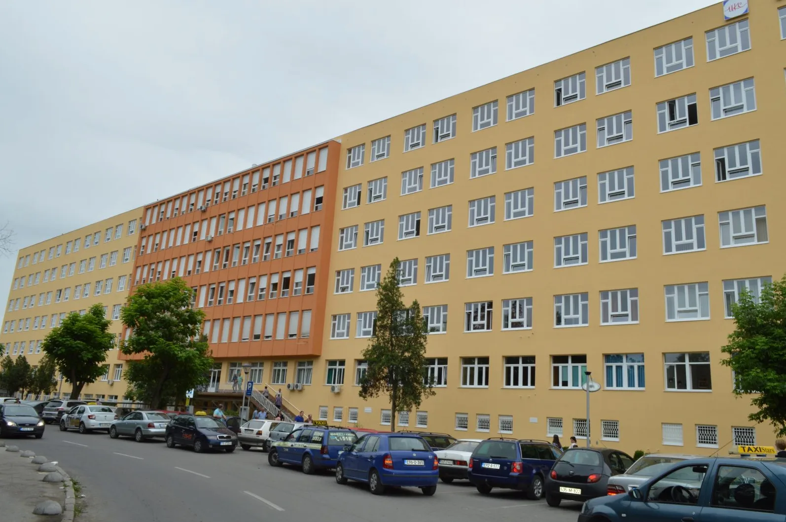 Žuta i narandžasta višespratnica sa brojnim prozorima, parkirani automobili i taksi vozila ispred.
