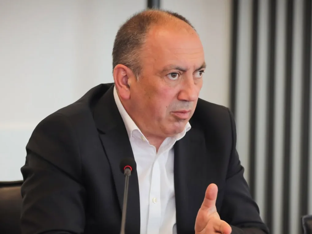 Biznismen u crnom odijelu i bijeloj košulji sjedi za stolom i govori ispred mikrofona.