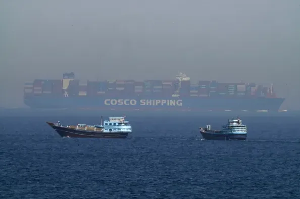 Dva manja drvena broda plove na pučini, dok se u daljini nazire ogroman COSCO Shipping kontejnerski brod.