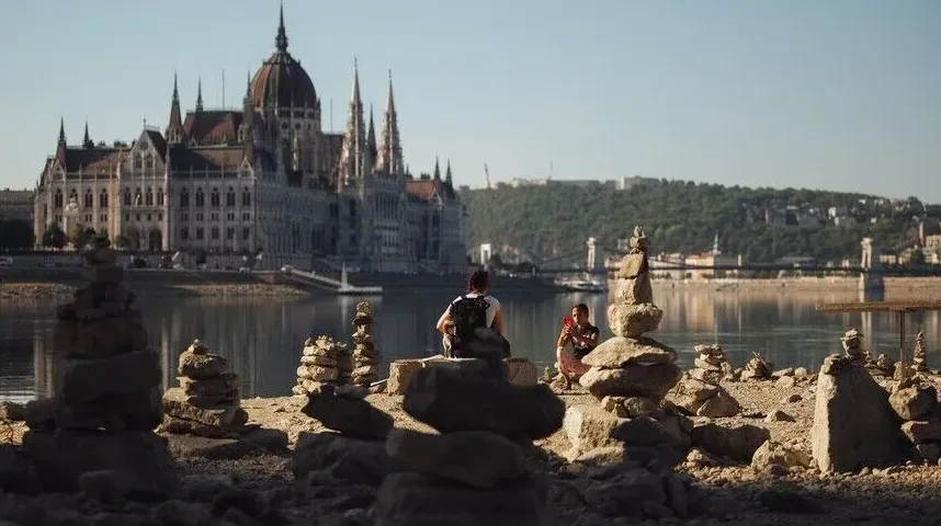 Osobe sjede pored Dunava, sa Budimpeštanskim parlamentom u pozadini, okruženi kamenim skulpturama.