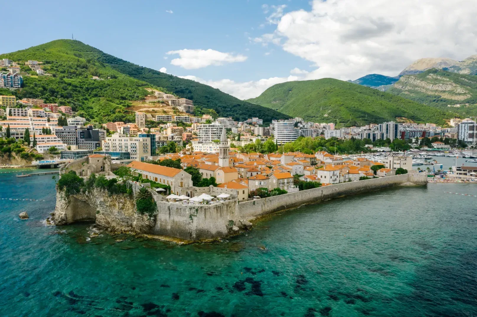 Aerialni pogled na Stari grad Budve i obalu sa zelenim brdima u pozadini.