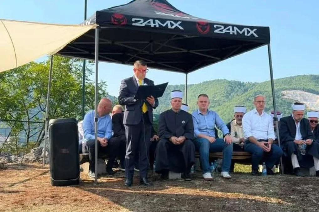 Grupa muškaraca, od kojih neki nose odijela, a neki islamske kape, sjede na klupi na otvorenom ispod šatora sa logom '24MX'. Jedan muškarac u odijelu stoji i drži dokument.