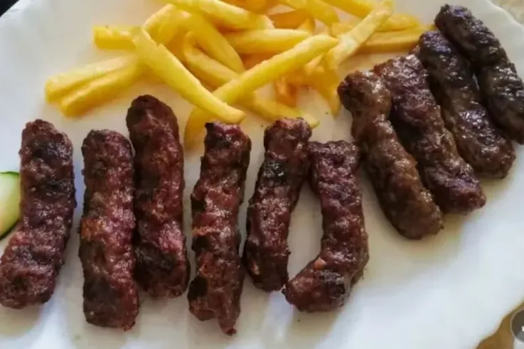 Ćevapi posluženi na tanjiru sa pomfritom i kriškom krastavca.