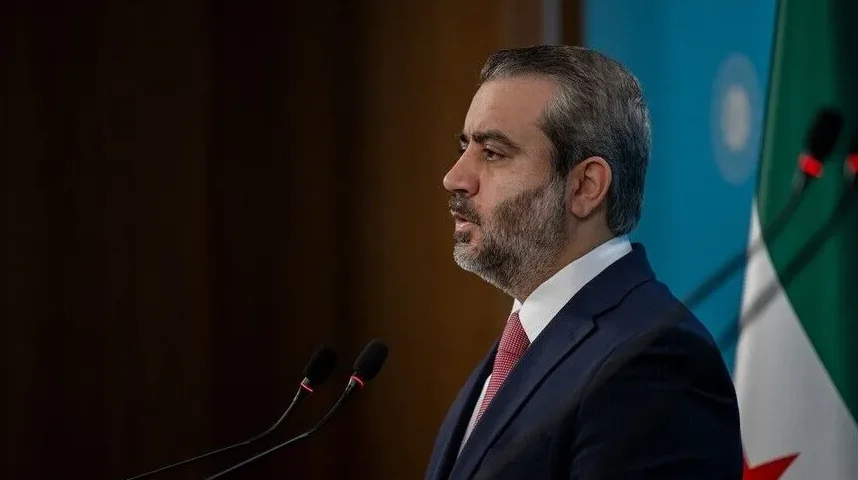Muškarac u tamnom odijelu i crvenoj kravati govori ispred podijuma, s dva mikrofona ispred sebe i zastavom u pozadini.