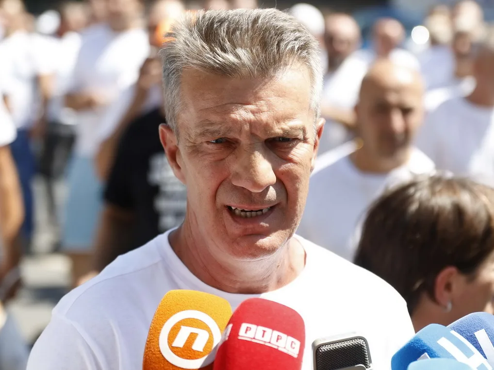Krupni kadar na lice starijeg muškarca sa kratkom sijedom kosom, koji govori u više mikrofona ispred sebe. Muškarac je odjeven u bijelu majicu. U pozadini se naziru brojne zamućene osobe, također odjevene u bijele majice.