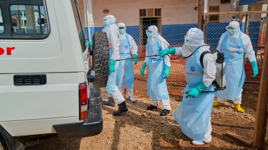 Lica u potpunoj zaštitnoj opremi za dezinfekciju, uključujući skafandere, maske, rukavice i vizire, provode dezinfekciju bijelog vozila i okolnog područja pomoću prskalica.