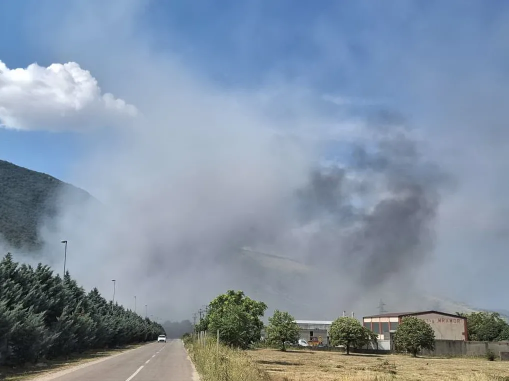Gusta bijela i crna dimna zavjesa širi se iznad industrijske zone i brdovitog krajolika pod vedrim nebom.