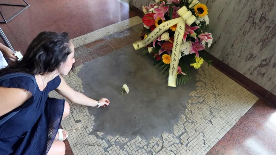 Djevojka u tamnoplavoj haljini kleči i polaže bijeli cvijet na spomen-obilježje, pored vijenca s natpisom.