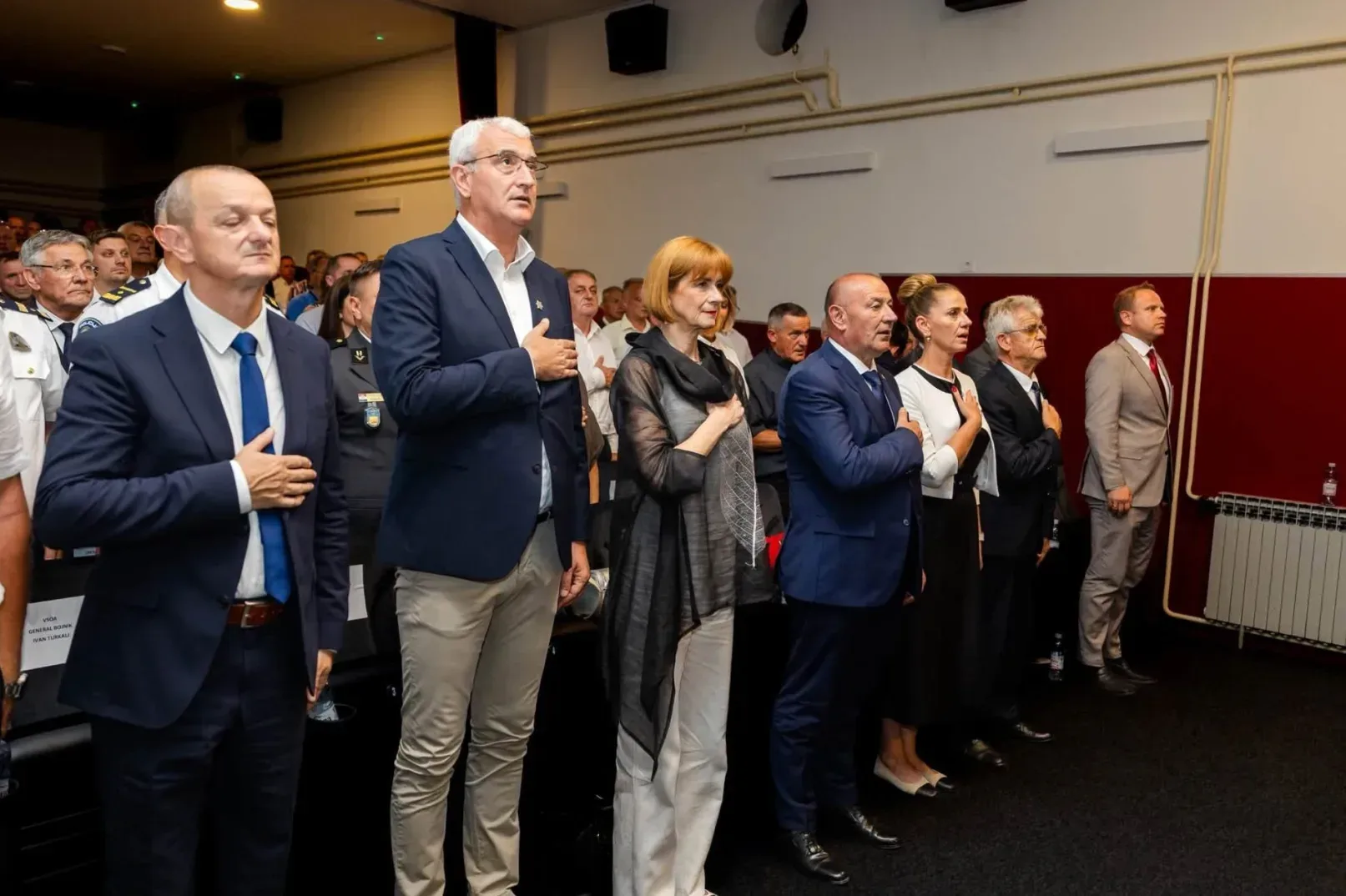Grupa ljudi u svečanim odijelima stoji u dvorani, sa rukama na srcu, vjerovatno tokom himne.