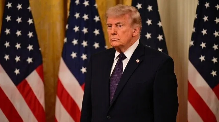 Donald Trump u tamnom odijelu i ljubičastoj kravati stoji ispred tri američke zastave.