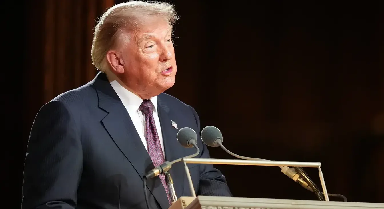 Donald Trump u tamnom odijelu i sa crvenom kravatom govori na bini sa dva mikrofona.