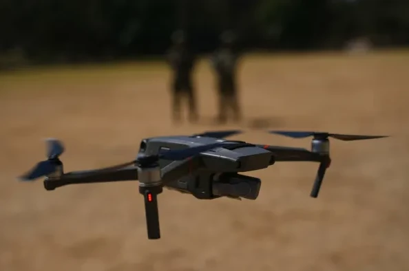 Moderna siva bespilotna letjelica (dron) sa crvenim LED svjetlom na jednom od kraka, u letu iznad pješčanog terena. U zamućenoj pozadini naziru se siluete dvije osobe u uniformama.