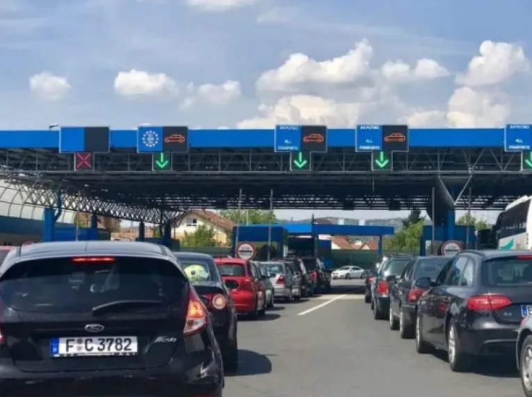 Duga kolona automobila čeka na graničnom prelazu, saobraćajni znaci pokazuju zelena svjetla za prolazak putničkih vozila i sve putnike.