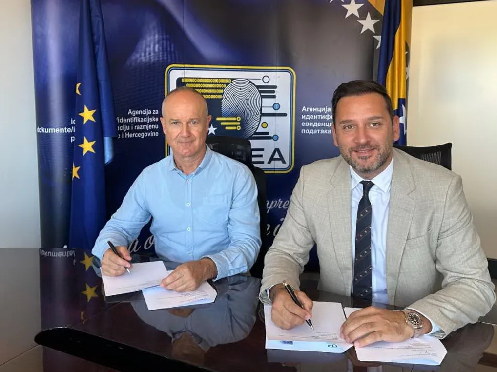Dvojica muškaraca sjede za stolom i potpisuju dokumente, sa zastavama Bosne i Hercegovine i Evropske unije u pozadini.