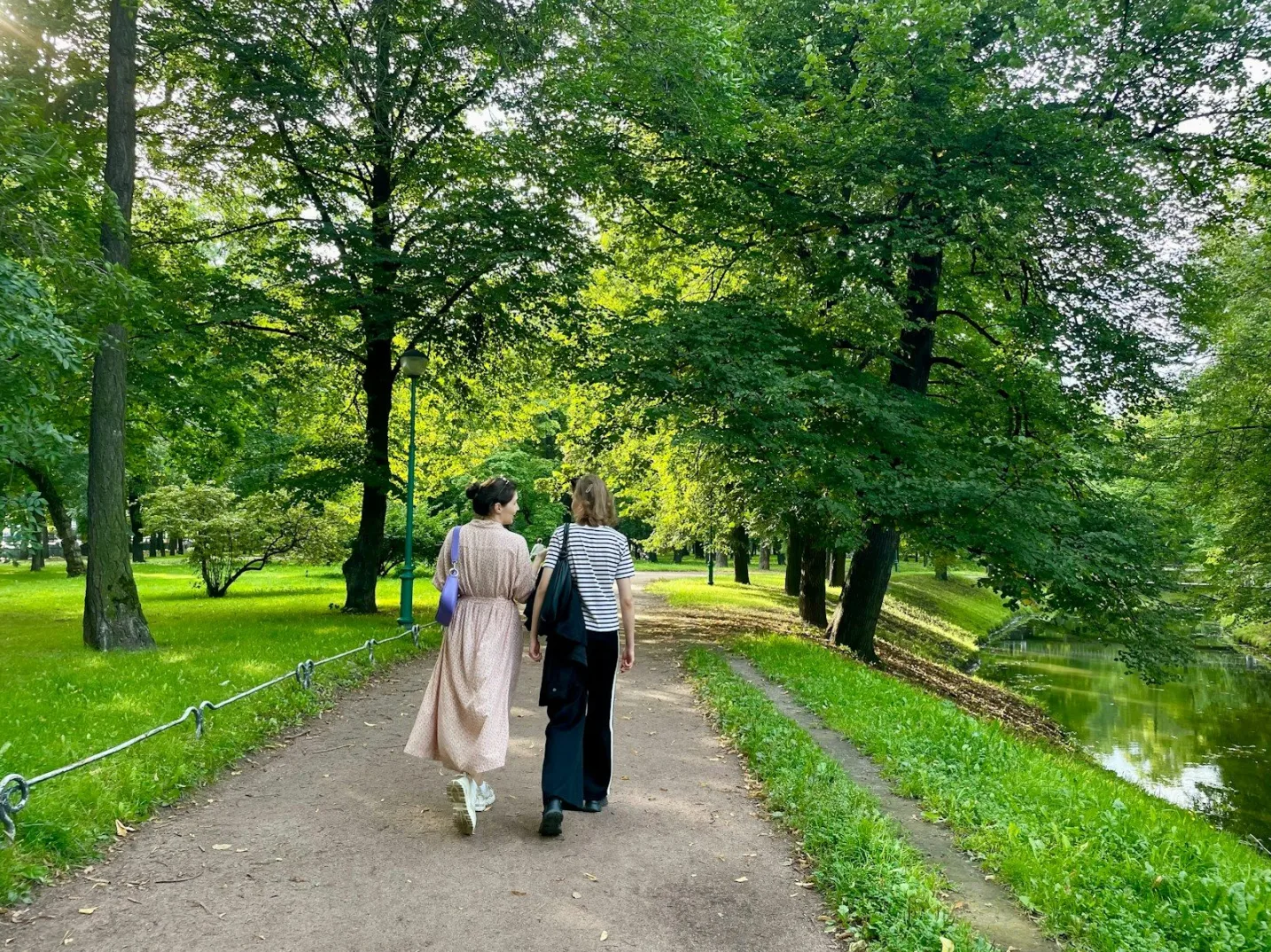 Dvije djevojke šetaju utabanom stazom kroz bujni zeleni park, okružene drvećem i travnjacima, dok se s desne strane nazire mirna rijeka.