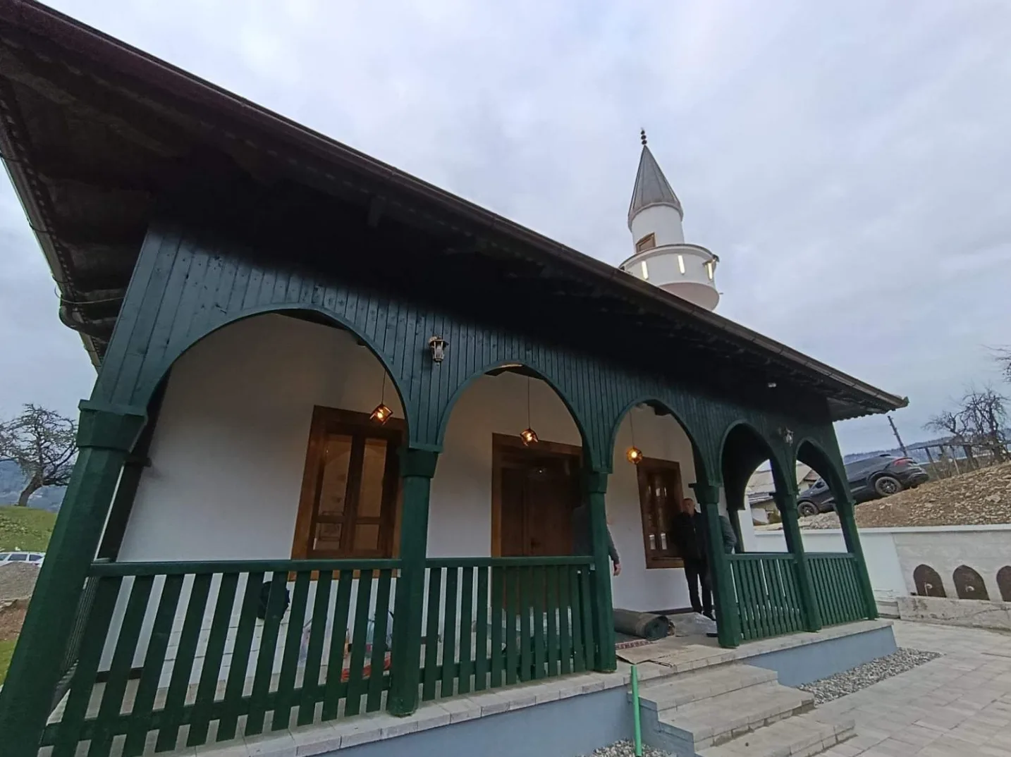 Fasada džamije sa zelenim drvenim trijemom, arkadama i minaretom u pozadini.