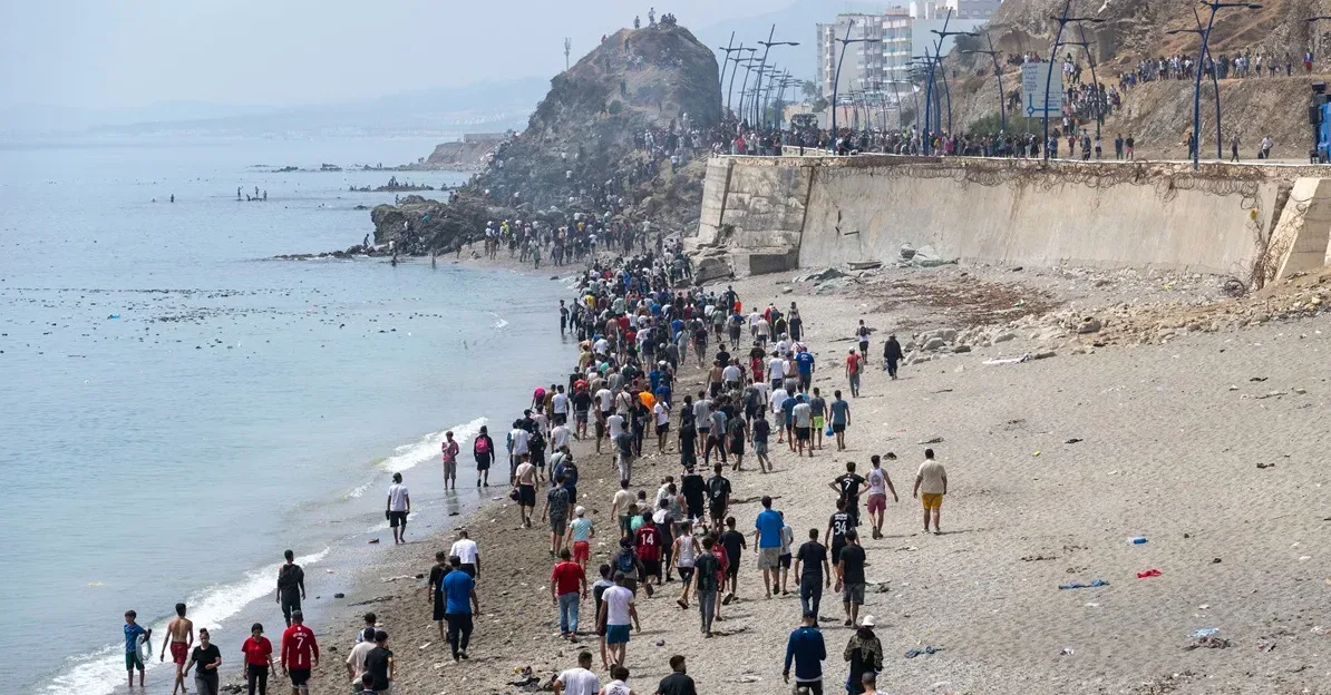 Velika grupa migranata pješice ide obalom mora, neki od njih su u vodi, a iza njih se vidi brdo sa zgradama u daljini.