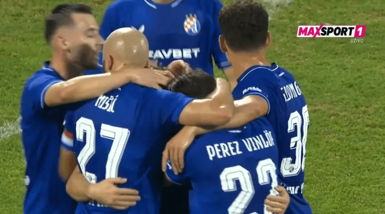 Fudbaleri u plavim dresovima grle se na terenu nakon postignutog gola, a na dresovima se vide brojevi i prezimena igrača.