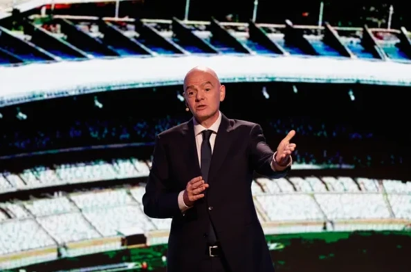 Đani Infantino u crnom odijelu sa bijelom košuljom i tamnom kravatom, drži ruke u gestikulaciji dok govori. U pozadini je prikaz stadiona.