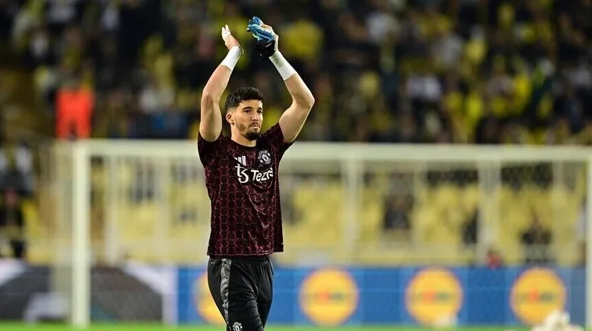 Fudbaler, golman Manchester Uniteda, sa podignutim rukama i rukavicama, odjeven u crveno-crnu garnituru, pozdravlja publiku na stadionu.