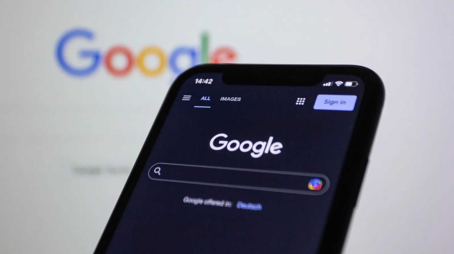 Mobilni telefon prikazuje Google pretraživač sa upaljenim ekranom, dok se u pozadini vidi logotip Googlea na desktop računaru.