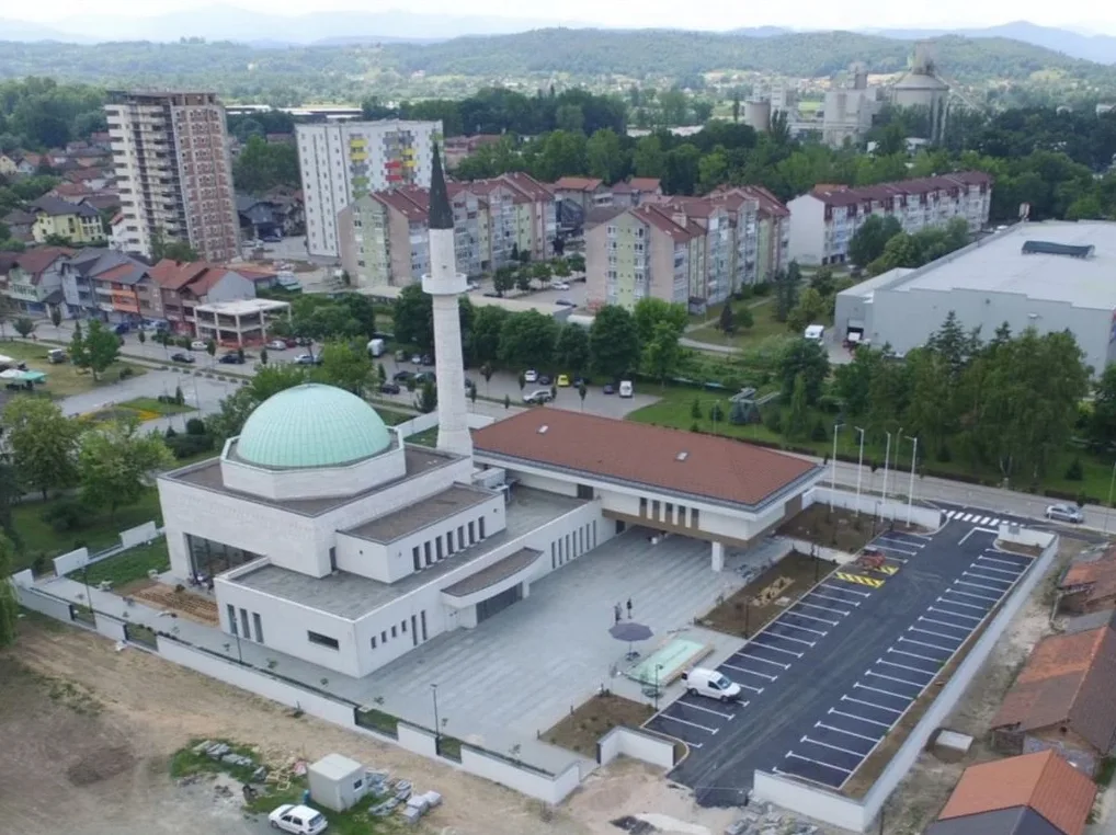 Pogled iz zraka na modernu gradsku džamiju sa zelenom kupolom i minaretom, okruženu stambenim zgradama, parkingom i urbanim pejzažem.