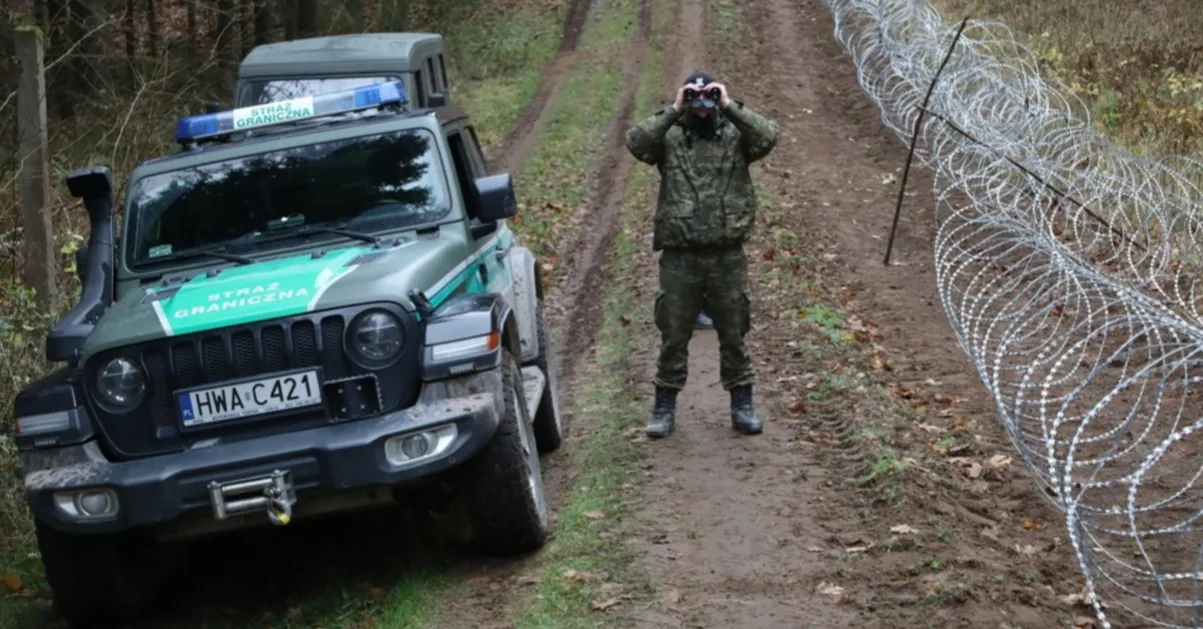 Granični službenik u uniformi posmatra kroz dalekozor na šumskom putu pored terenskog vozila sa oznakom "STRAŻ GRANICZNA" i ograde od bodljikave žice.