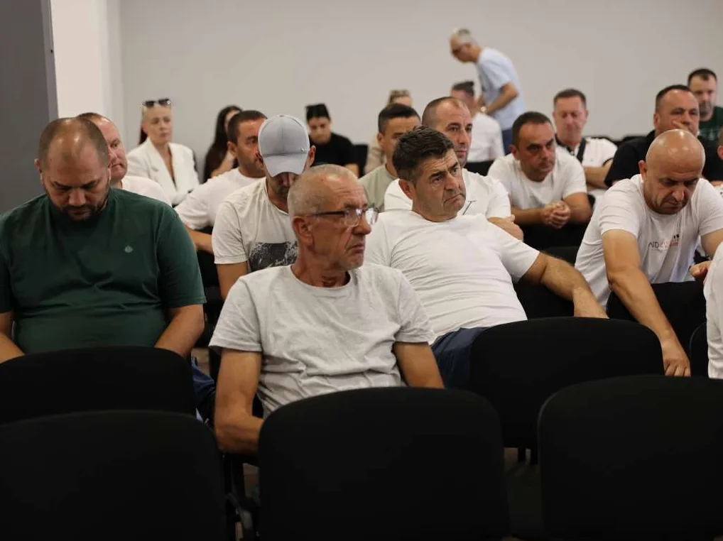 Grupa muškaraca sjedi u gledalištu, neki nose bele majice, a jedan nosi zelenu majicu.