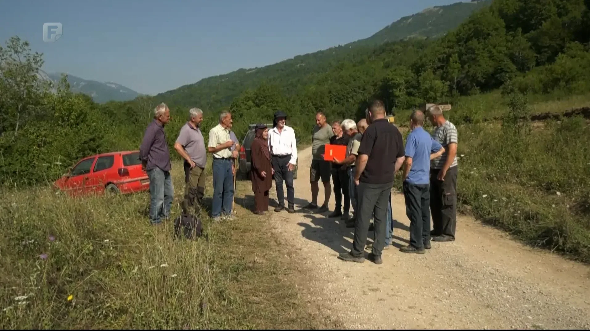 Grupa muškaraca i jedna žena okupljeni su na šljunčanoj stazi usred zelenila i šume. U pozadini se vidi crveni automobil koji je djelomično sišao s puta. Neki od muškaraca nose odjeću za rad na otvorenom, a jedan od njih drži crveni dokument. Izgleda kao da se raspravlja o nečemu ili da se organizuje radna akcija.