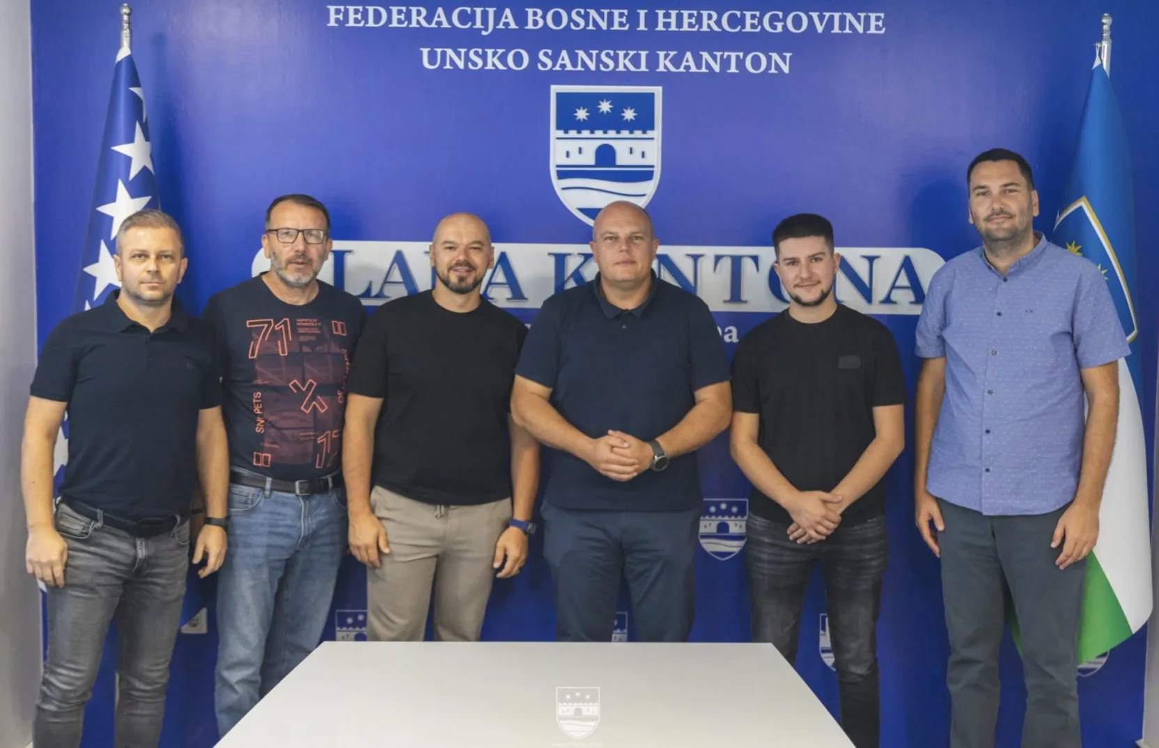 Šest muškaraca u poluportretu stoji u redu ispred plave pozadine sa grbom Unsko-sanskog kantona i zastavom Bosne i Hercegovine.