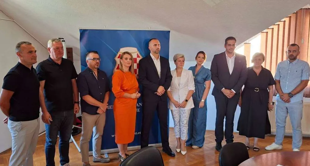 Grupa od devet osoba, muškaraca i žena, stoji ispred plavog panoa s grbom na kojem piše nešto na bosanskom jeziku.