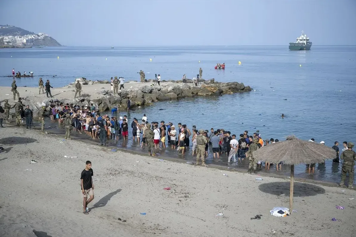 Skupina migranata stoji u vodi na obali, okružena pripadnicima vojske i policije. U pozadini se vidi more, brod i obalni grad.
