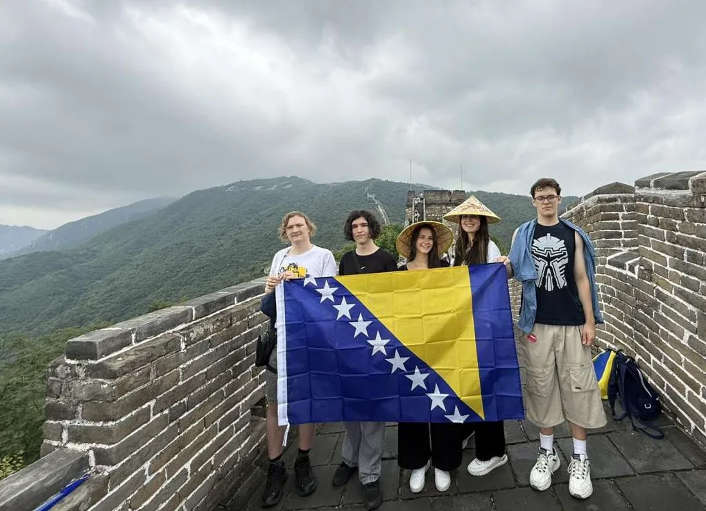 Grupa mladih ljudi drži zastavu Bosne i Hercegovine na vrhu Kineskog zida, s planinskim pejzažem u pozadini.