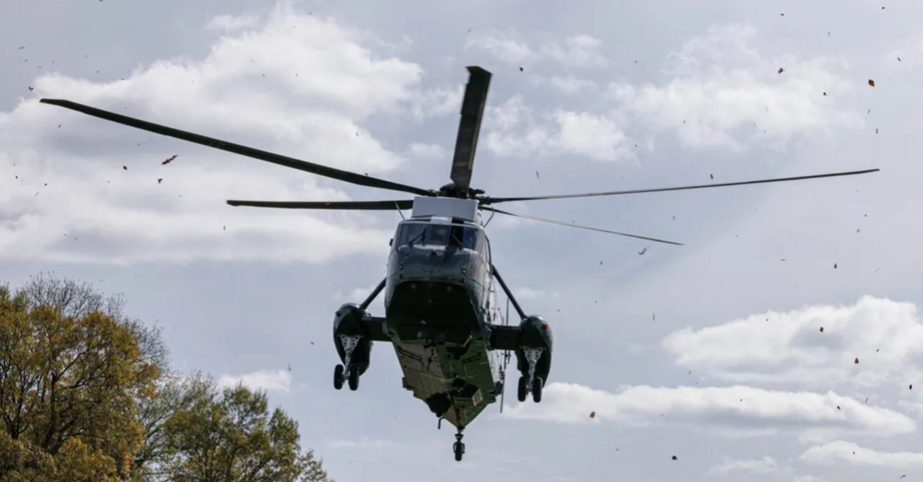 Veliki tamnozeleni helikopter, sličan američkom Marine One, u niskom letu iznad drveća s jesenjim lišćem koje vjetar nosi kroz nebo.